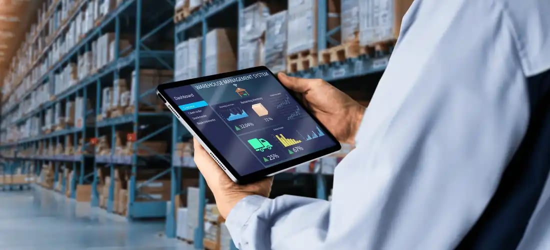 Warehouse manager using a tablet with a digital warehouse management system (WMS) dashboard to track inventory, shipments, and stock levels in real-time. The background shows an organized warehouse with shelves stocked with goods, representing smart logistics and Manufacturing 4.0 efficiency.